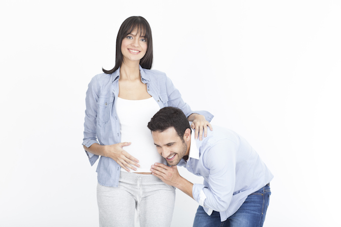 Dad bending over playfully to listen to wife’s pregnant belly