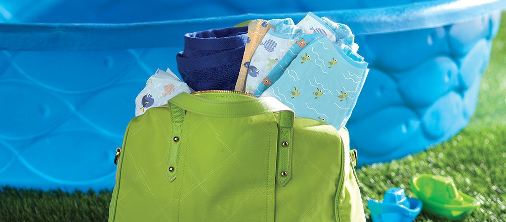 Una bolsa de pañales verde llena de calzoncitos Huggies Little Swimmers con variados diseños de Buscando a Nemo junto a una piscina para bebés