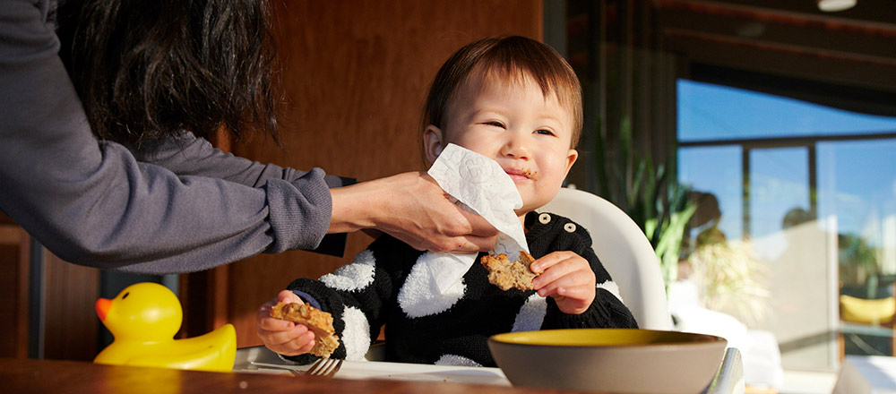 Una madre se inclina para limpiar la cara sucia de su bebé mientras come en una silla alta