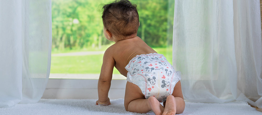 A diapered baby looking out a window on a sunny day
