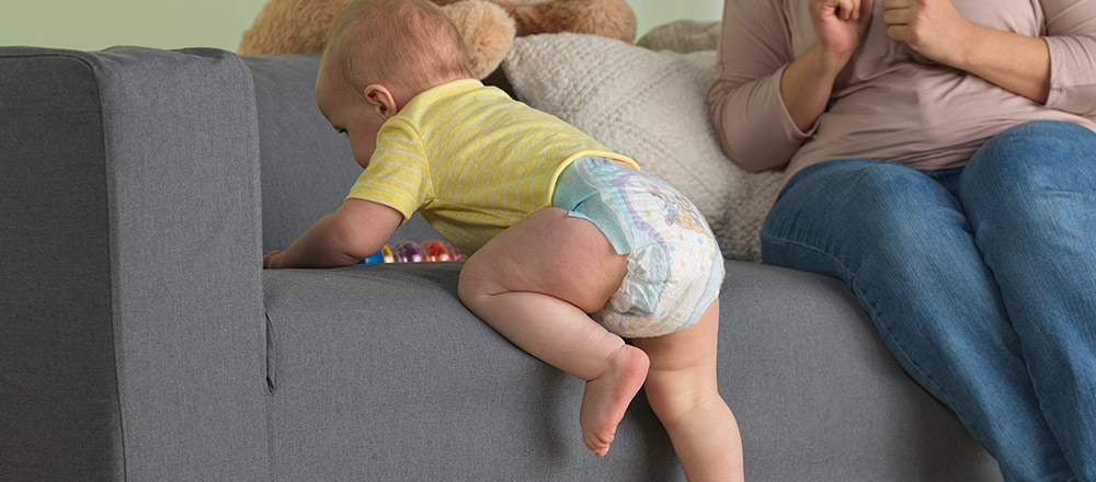 Un bebé con un pañal Huggies Little Movers Slip-On sube a un sofá junto a su madre