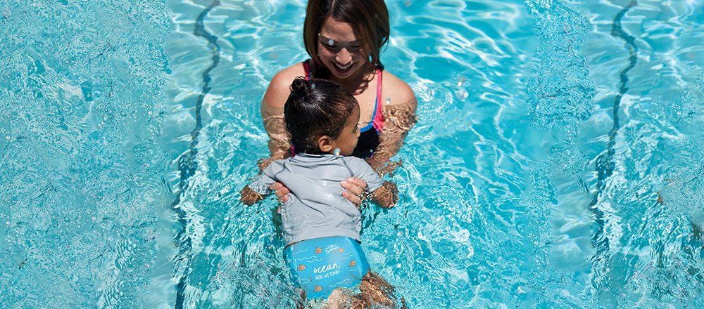 A laughing mother holds her baby wearing a pair of Huggies Little Swimmers Swim Pants in a pool