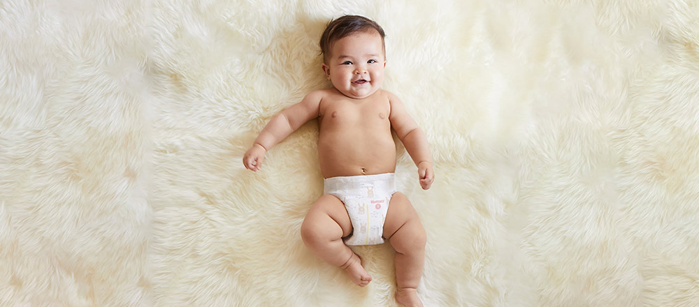 A diapered baby smiles while laying on a fuzzy of-white blanket