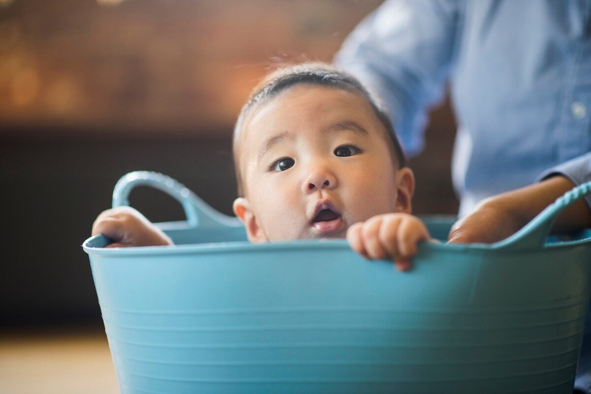 A baby sitting in a blue tote peers out curiously