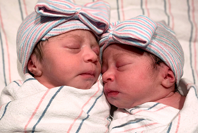 Newborn twins, Arya and Alessia, lay swaddled in matching outfits while cuddling each other