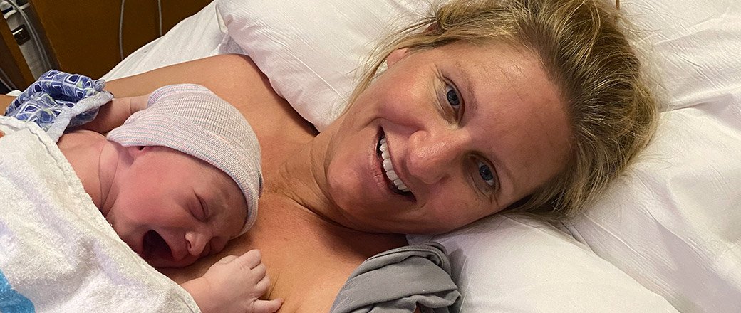 A smiling mother lays on a hospital bed with her crying newborn lying on her chest