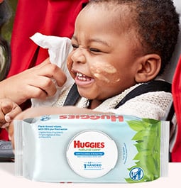 Una mano se acerca para limpiar con una toallita Huggies Natural Care la cara desordenada de un bebé que ríe