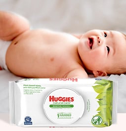 Un bebé en pañales se ríe mientras está acostado, detrás de una imagen de toallitas Huggies Natural Care