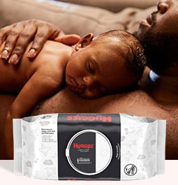 A father lays on a couch while his baby lays on his chest behind an image of Huggies Special Delivery Wipes