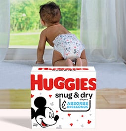 Un bébé portant une couche regarde par la fenêtre en une journée ensoleillée derrière une image d&#39;une boîte de couches Snug&nbsp;&amp;&nbsp;Dry de Huggies