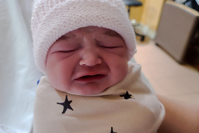 Newborn baby, Khloe, cries while swaddled in a white blanket and a white beanie