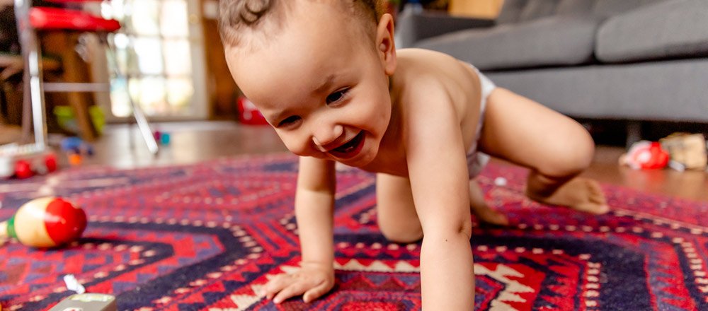 A laughing baby wearing a Huggies Little Movers Plus Diaper crawls on a rug