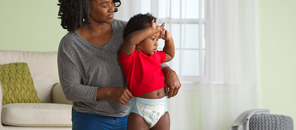 A smiling mother pulls a pair of Huggies Little Movers Slip-On Diapers onto her standing baby