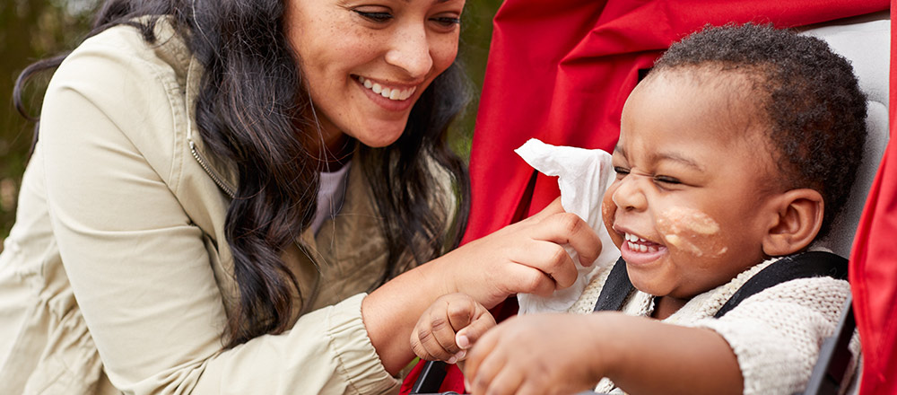 Una madre sonriente se acerca para limpiar con una toallita refrescante Huggies Natural Care la cara desordenada de un bebé que ríe