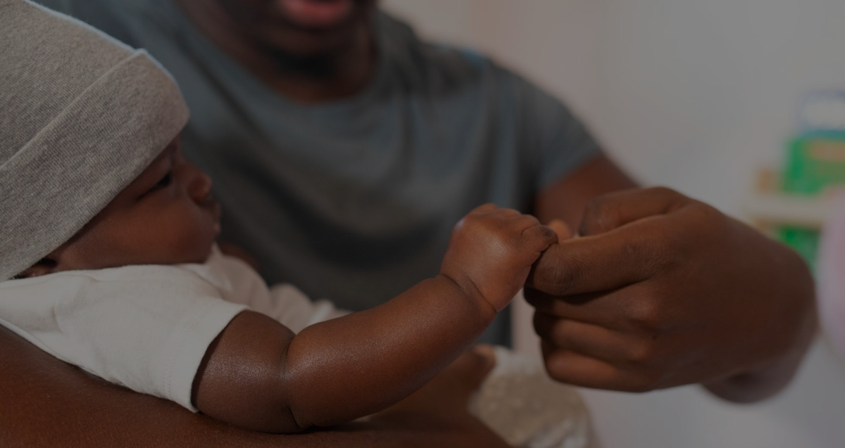 Un bebé con un gorro gris y una camisa blanca sujetando el dedo de su papá
