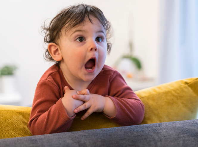 A surprised baby with folded hands leans on a yellow cushion