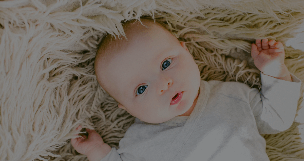 Un bebé acostado sobre una manta beige peluda mirando hacia arriba con los brazos cerca de su cabeza