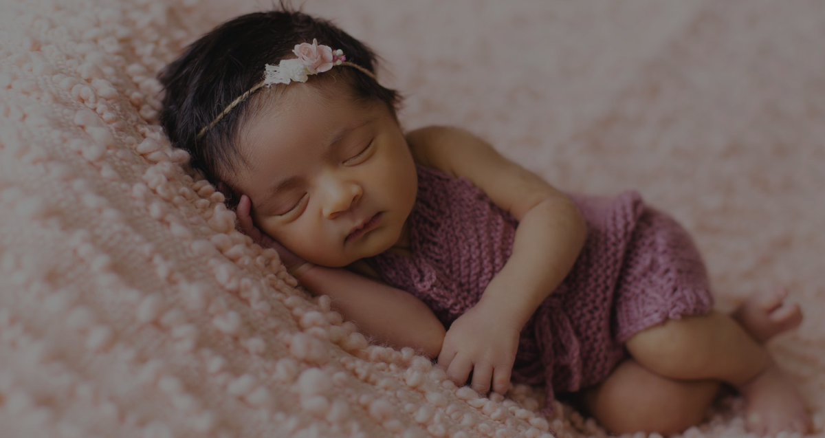 Baby wearing pink clothes lays on its side with eyes closed on a light pink blanket