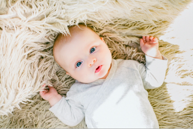 Baby laying on a beige fuzzy blanket looking up with arms near their head