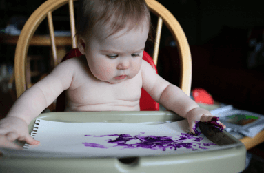 A baby sits in a high chair using purple finger paints on paper