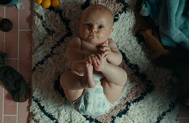 A diapered baby laying on a shag rug holds its feet