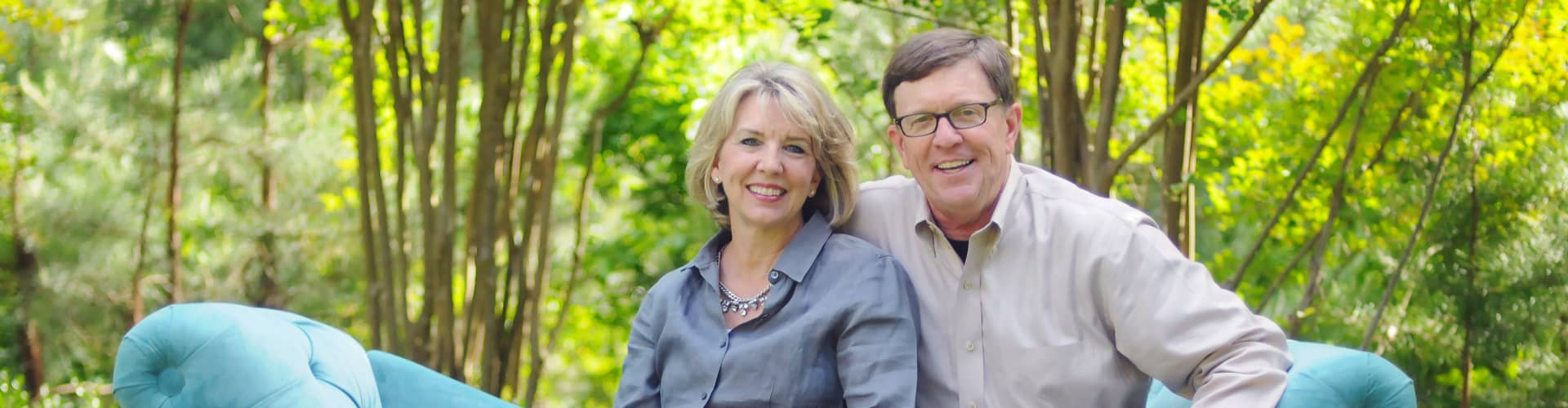 Dennis and Barbara Rainey smiling, sitting on a couch in the woods.